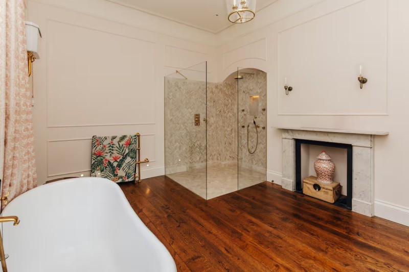 Luxurious bathroom with herringbone-tiled walk-in shower, freestanding bathtub with brass fixtures, floral towel radiator, wooden flooring, and a decorative fireplace with a ceramic vase.