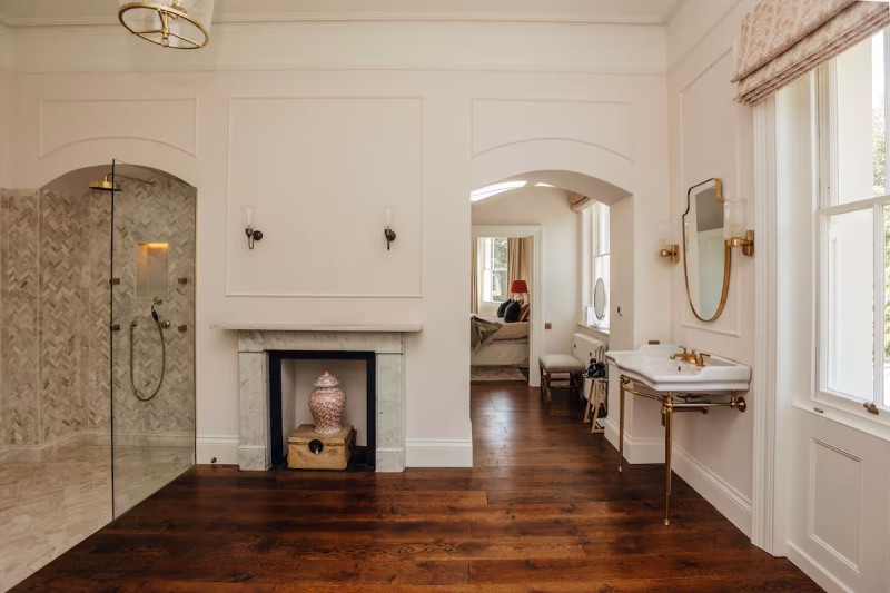 Elegant period-style bathroom with walk-in marble shower, vintage fireplace, and gold-accented basin leading into a luxury bedroom.