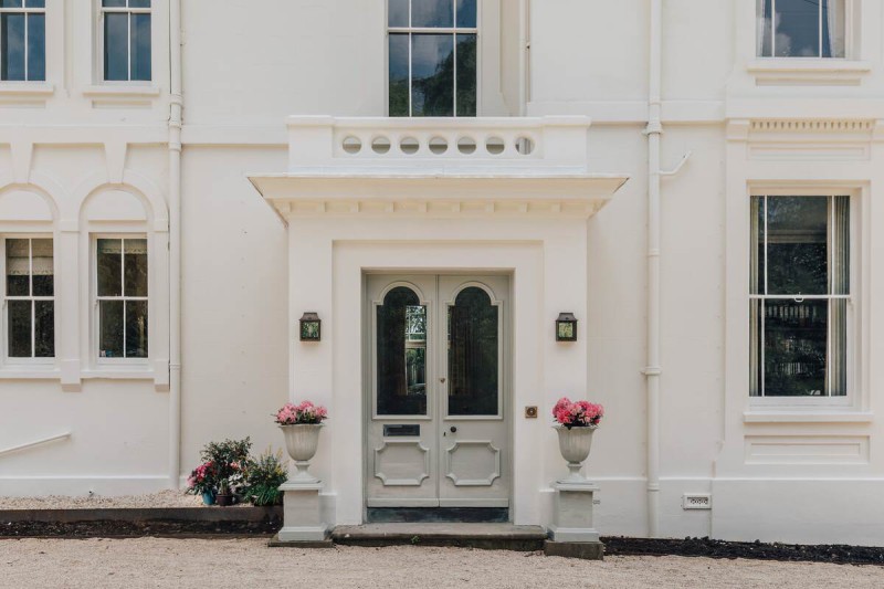 Elegant front entrance of a white-painted period house with a symmetrical double door, two wall lanterns, and pink flowers in classical urn planters on stone pedestals.