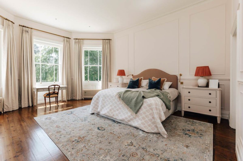 Bright and elegant master bedroom with large sash windows, classic wall panelling, and a soft neutral palette accented by statement lighting and rich wooden flooring.