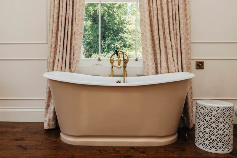Freestanding blush bathtub with vintage gold taps, framed by floral curtains and set against a large window with garden views — elegant, spa-like bathroom design.