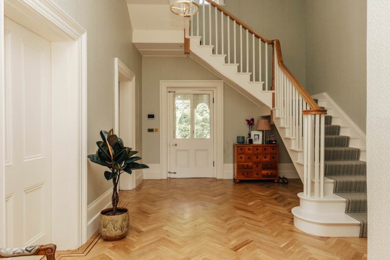 Elegant hallway with herringbone wood flooring, a curved staircase with white spindles and wooden handrail, a potted rubber plant, and a wooden chest of drawers beneath the stairs.