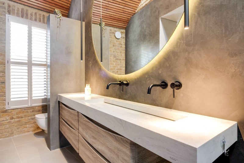 Modern bathroom with floating vanity, curved backlit mirror, black wall-mounted taps, and exposed brickwork