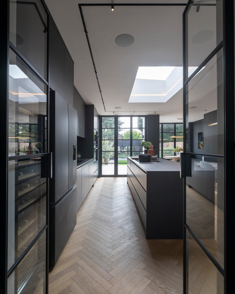 Sleek open-plan kitchen with black cabinetry, central island, herringbone flooring, and full-height glazing to garden