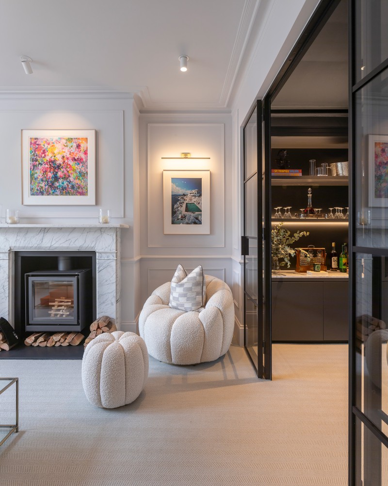 Elegant lounge with boucle armchair and pouffe, marble fireplace, and a view into a backlit modern bar nook through black-framed glass doors