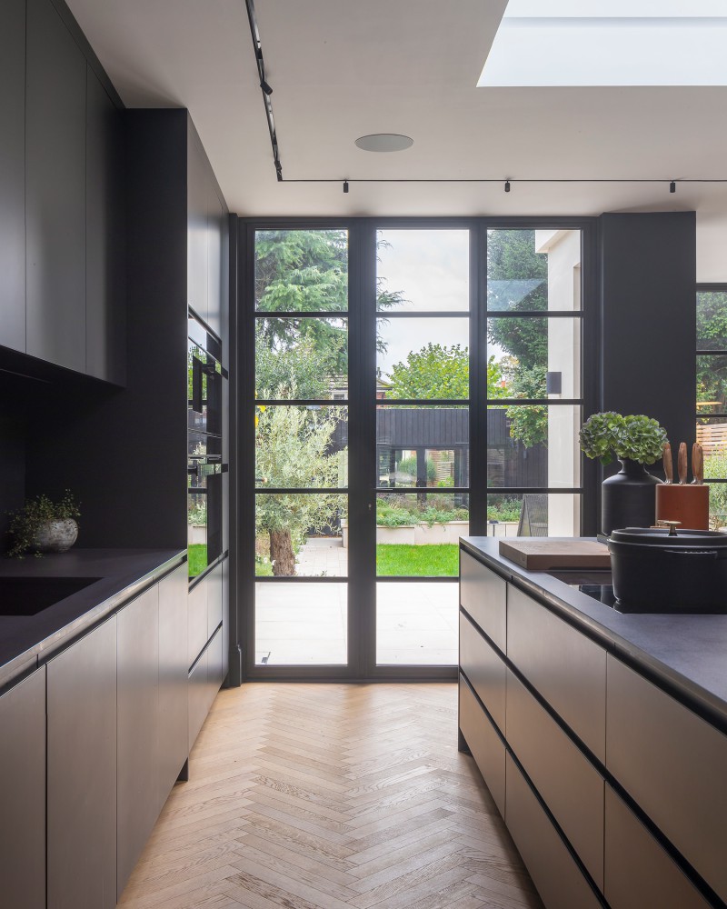 Modern kitchen with black handleless units, central island, herringbone wood flooring, and garden views through steel-framed doors